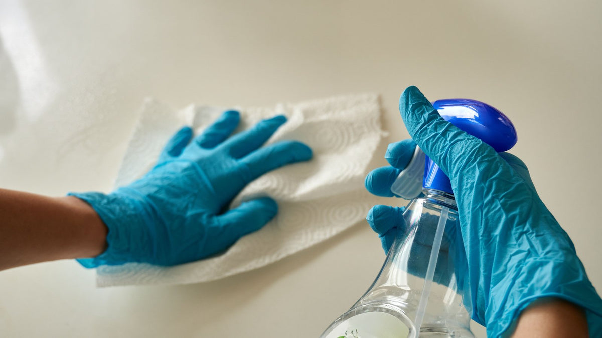 Gloved hands spraying cleaner and wiping a surface with a paper towel