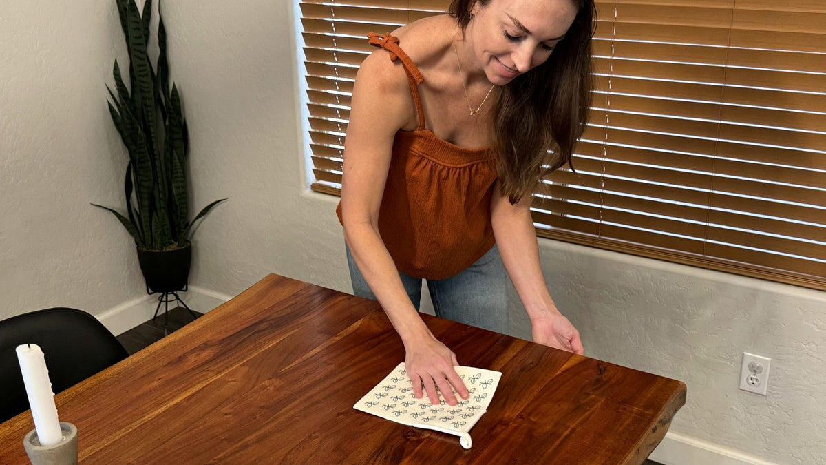 Woman wiping a sealed walnut-toned wood dining table with a damp Giro Swedish dishcloth