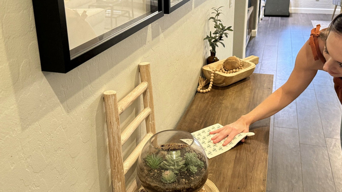 Woman dusting a wooden console table with a Swedish dishcloth beside a succulent terrarium
