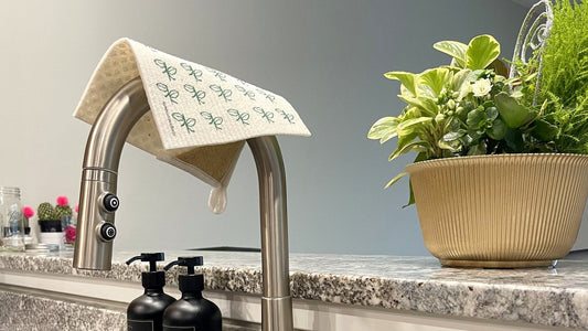 Bright kitchen sink with a Swedish dishcloth draped over the faucet, soap bottles and plants