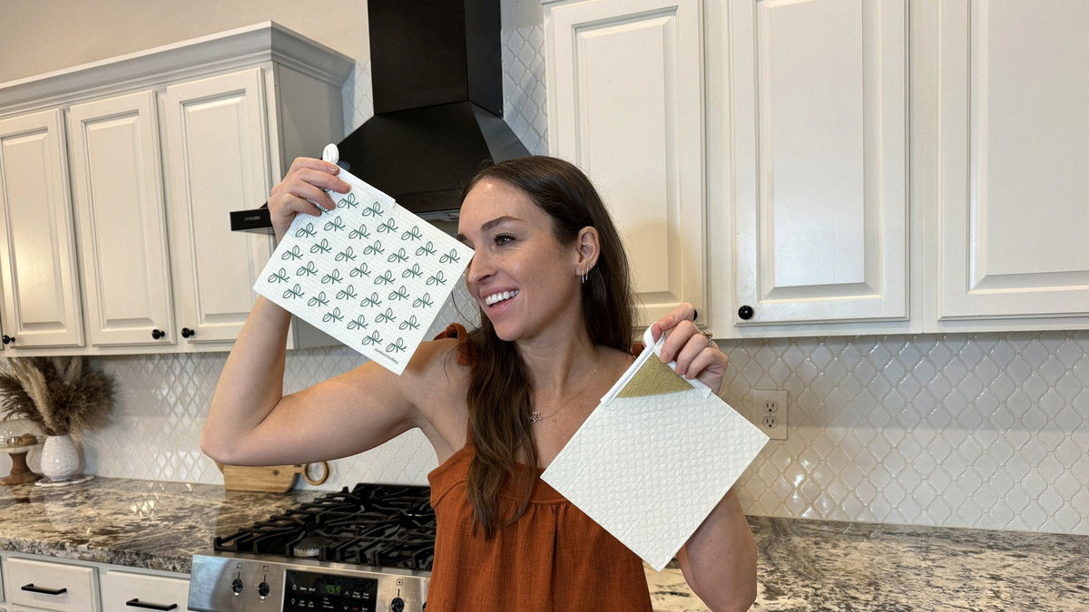 Smiling woman holding two Giro Swedish dishcloths in a bright kitchen